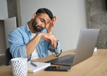 Young indian eastern tired exhausted business man rubbing eyes sitting in modern home office with laptop on desk. Overworked burnout academic Hispanic student with glasses in hand feeling eyestrain.