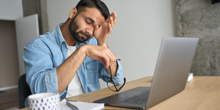 Young indian eastern tired exhausted business man rubbing eyes sitting in modern home office with laptop on desk. Overworked burnout academic Hispanic student with glasses in hand feeling eyestrain.