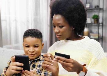 Close up of beautiful young african american mom sitting on sofa with son holding bank card and smartphone together shopping online.