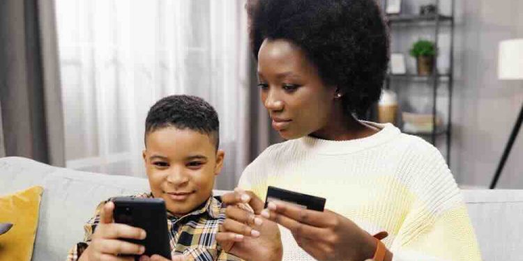 Close up of beautiful young african american mom sitting on sofa with son holding bank card and smartphone together shopping online.