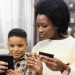 Close up of beautiful young african american mom sitting on sofa with son holding bank card and smartphone together shopping online.