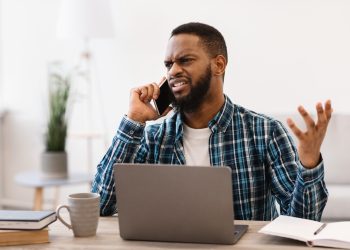 Displeased Black Business Guy Talking On Phone Having Quarrel By Cellphone Sitting At Workplace In Modern Office. Angry Customer Calling To Hotline Having Problem With Computer