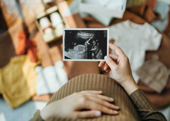 Personal perspective of Asian pregnant woman looking at an ultrasound scan photo while gently touching her baby bump, with baby clothings and accessories laying on the floor. Mother-to-be. Preparation for a new family member. Expecting a new life concept