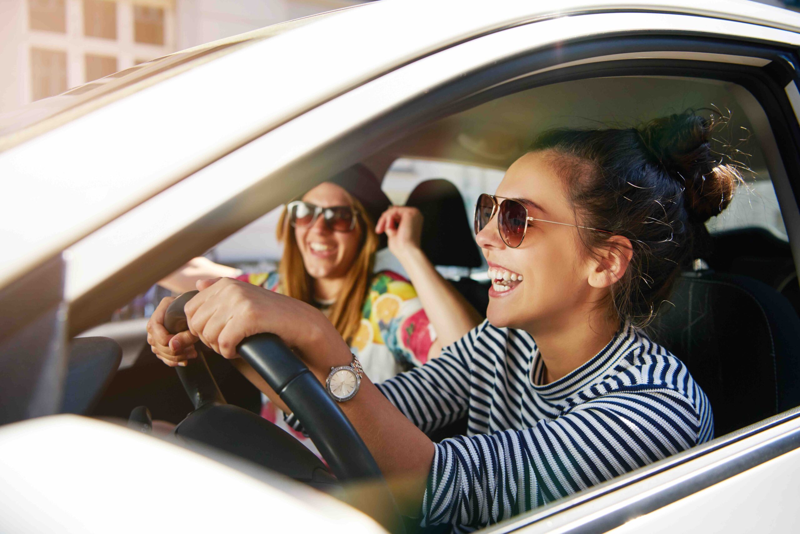 Laughing trendy young girlfriends in sunglasses traveling in a car in town with focus to the profile of an attractive young woman through the open window