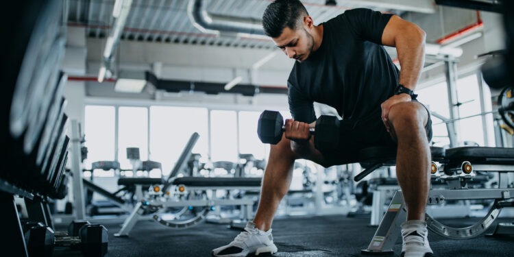Man sitting on one gym machine and doing biceps.