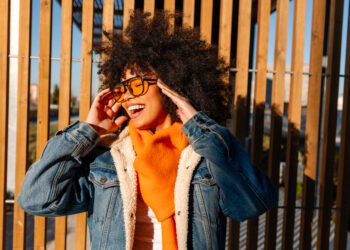 Stylish young woman with afro hair, wearing orange scarf and denim jacket, smiling and adjusting sunglasses against wooden background