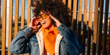 Stylish young woman with afro hair, wearing orange scarf and denim jacket, smiling and adjusting sunglasses against wooden background