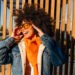 Stylish young woman with afro hair, wearing orange scarf and denim jacket, smiling and adjusting sunglasses against wooden background