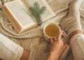 Cozy winter evening , warm woolen socks. Woman is lying feet up on white shaggy blanket and reading book. Cozy leisure scene. Text in book is unreadable. Woman relaxing at home. Comfy lifestyle.