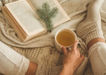 Cozy winter evening , warm woolen socks. Woman is lying feet up on white shaggy blanket and reading book. Cozy leisure scene. Text in book is unreadable. Woman relaxing at home. Comfy lifestyle.