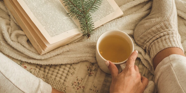 Cozy winter evening , warm woolen socks. Woman is lying feet up on white shaggy blanket and reading book. Cozy leisure scene. Text in book is unreadable. Woman relaxing at home. Comfy lifestyle.