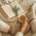 Cozy winter evening , warm woolen socks. Woman is lying feet up on white shaggy blanket and reading book. Cozy leisure scene. Text in book is unreadable. Woman relaxing at home. Comfy lifestyle.