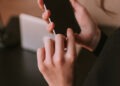 Woman;s hand holding cellphone ready to unlocked it. Girl using telephone near table wit computer in office